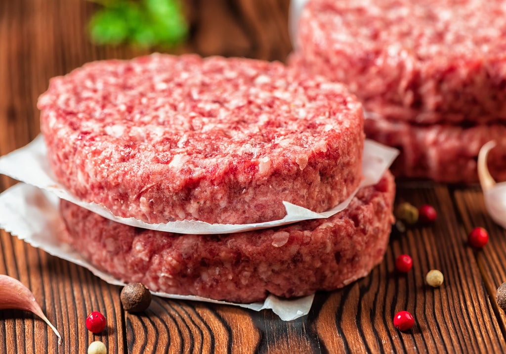 Raw beef burgers displayed by pairs. Peppercorn and garlic garnish the cutting board. King George's Steaks and Seafood.