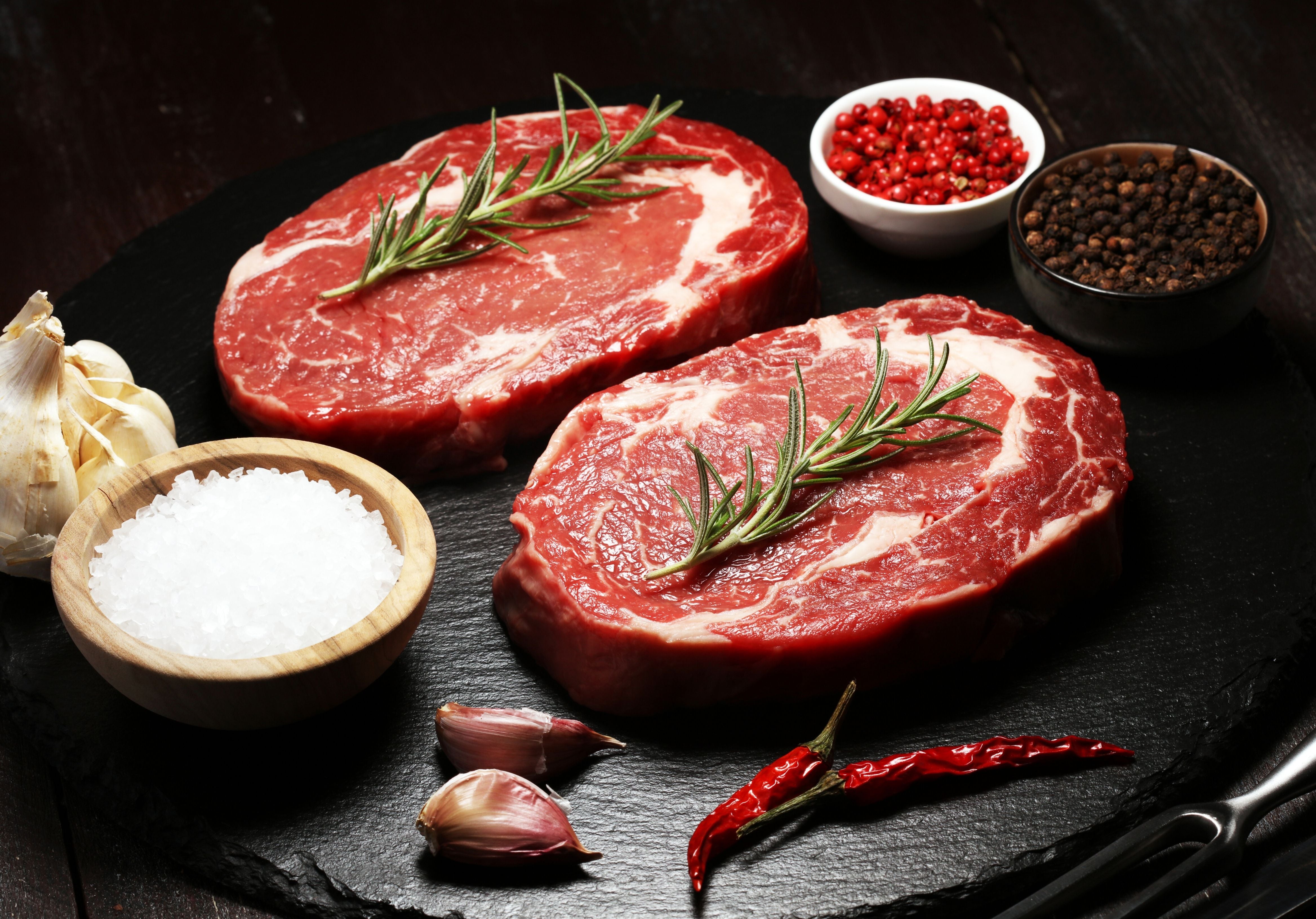 Two Rib Eye Prime fillets displayed on a cutting board displayed with sea salt, garlic, dry red chili, peppercorn, and rosemary. King George's Steaks and Seafood.