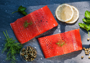 Two raw Sockeye Salmon fillets displayed with fresh capers, dill, coriander leaves,, lemon wheels, peppercorn and sea salt. King George's Steaks and Seafood.