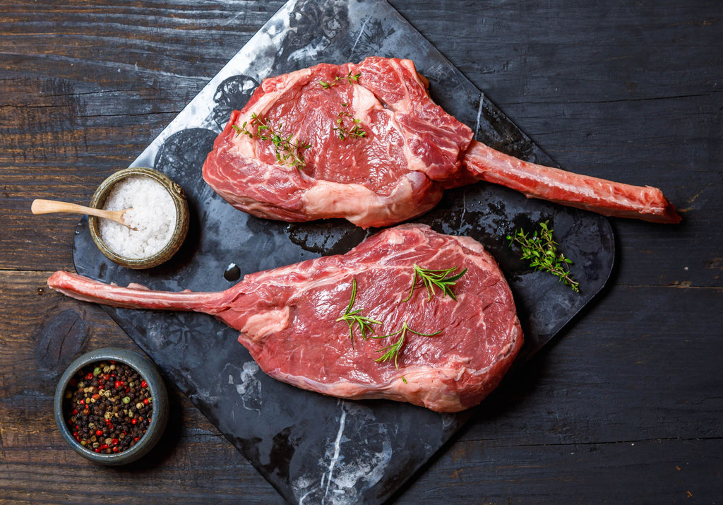 Two raw Tomahawk Steaks on a cutting board, garnished with rosemary, peppercorn and sea salt. King George's Steak and Seafood. 